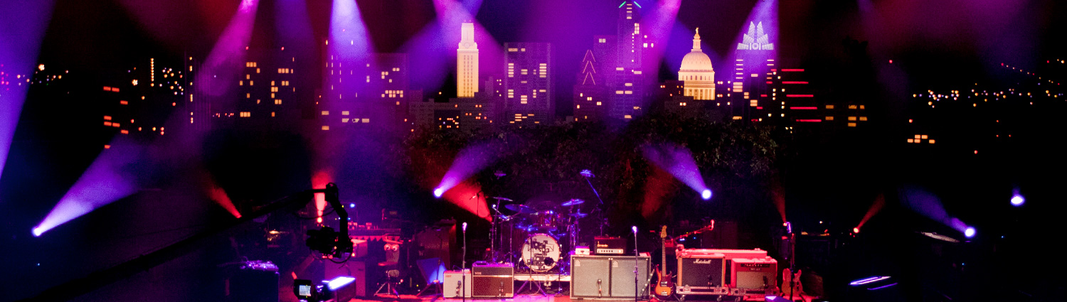 The Austin City Limits stage, coverd with insturments like guitars, a drumset, and keyboards, lit up with stage lights, and the Austin City Limits backdrop.
